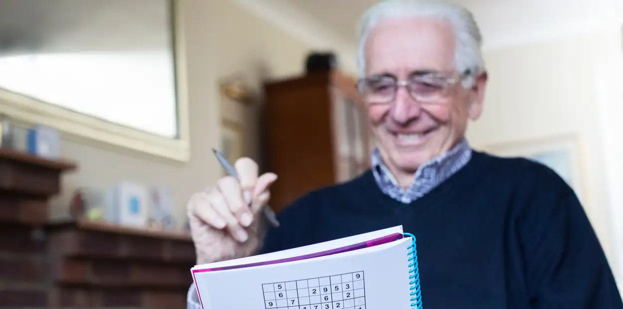 A senior man sitting in his home at Monarch Landing in IL and working on a sudoku puzzle, one of the best brain games for seniors.